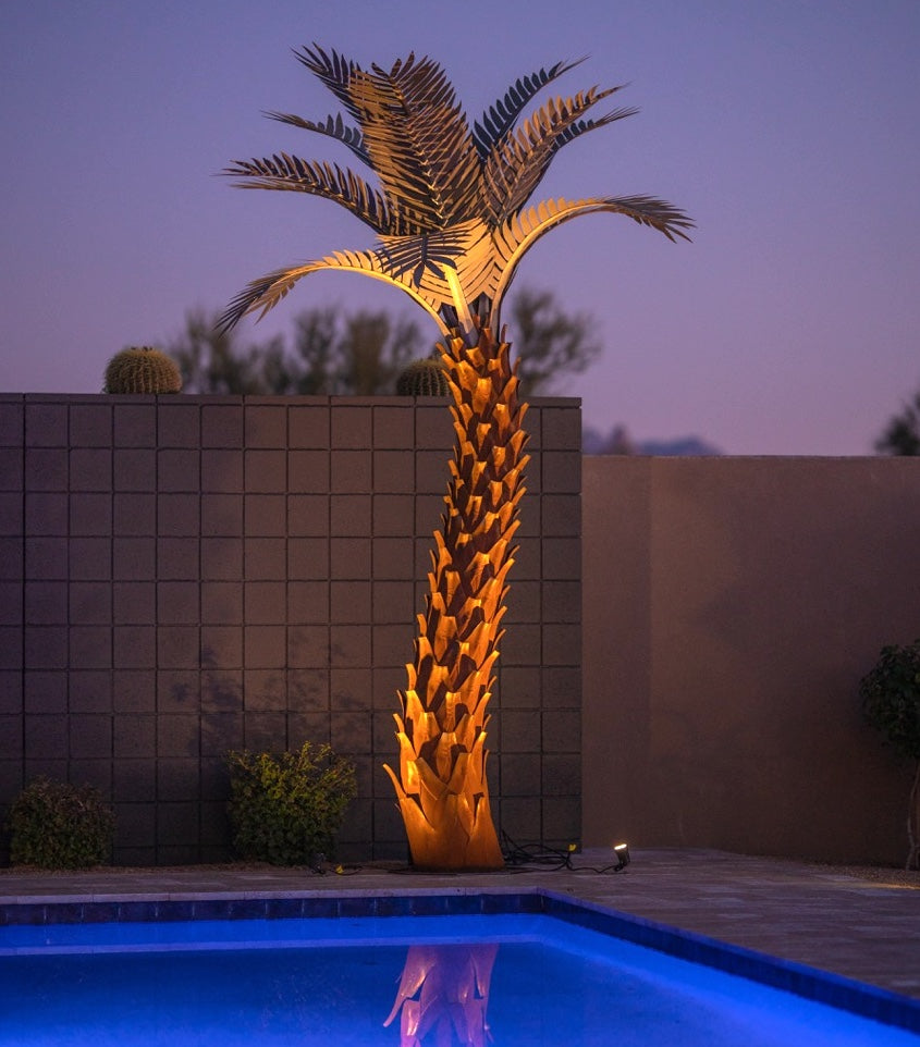 A large metal sunset palm tree that is illuminated, standing 10ft tall, located in a backyard near a pool. The sculpture's intricate design features realistic palm fronds and textured trunk, creating a lifelike appearance.