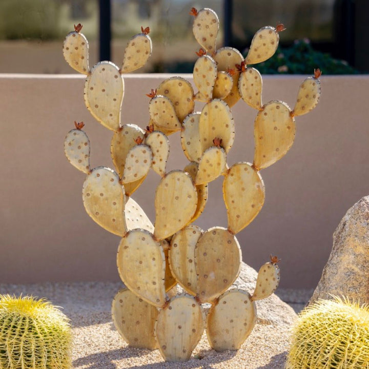 A handcrafted realistic large metal prickly pear cactus planted into gravel in a yard. Two real cactus' can be seen either side.