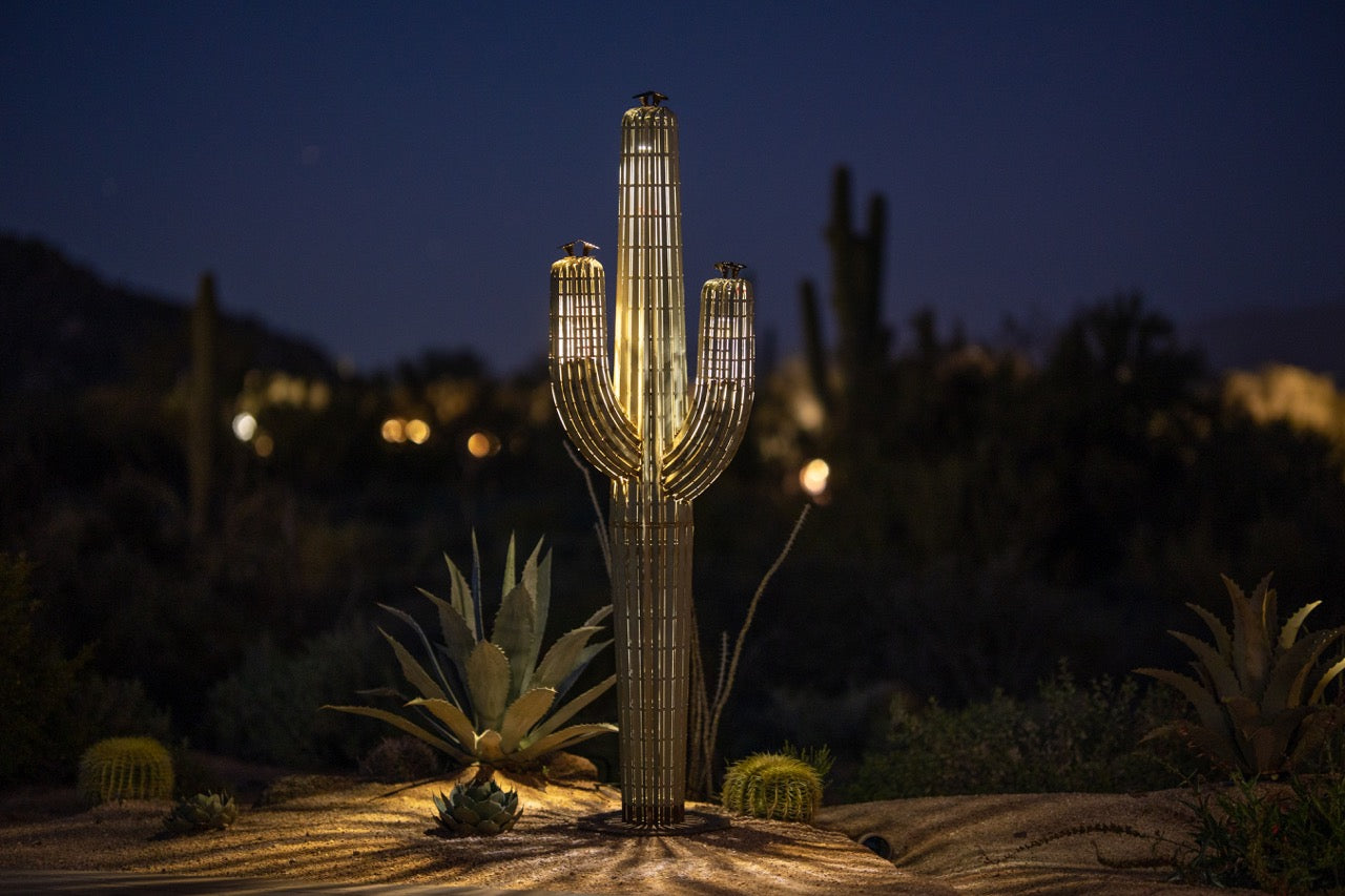 A large metal saguaro cactus illuminated at night in the desert. The sculpture's intricate design features a realistic saguaro body and arms, creating a lifelike appearance.