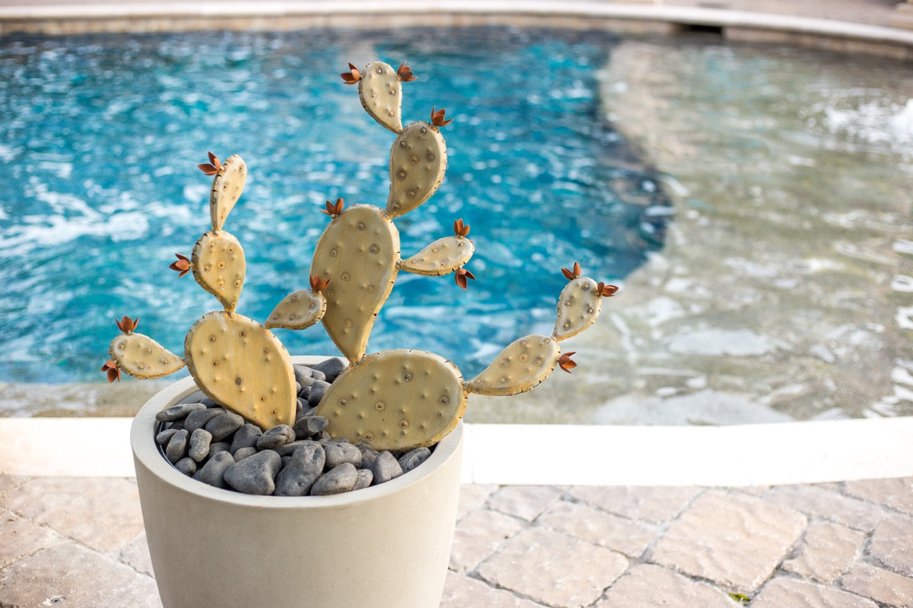 A handcrafted realistic metal prickly pear cactus in a white planter besides a pool in the back yard.
