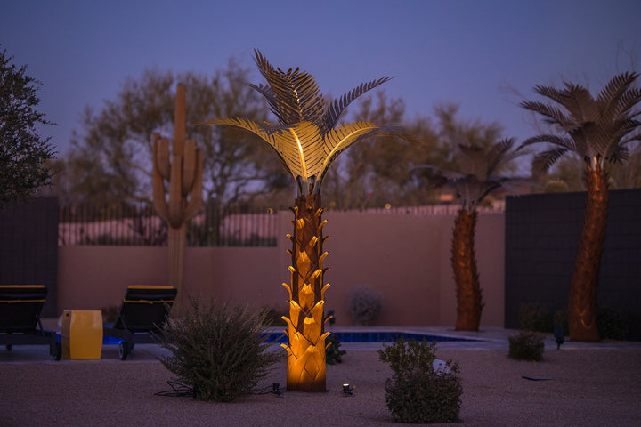 A large metal paradise palm tree that is illuminated, standing 10ft tall, located in a backyard near a pool. The sculpture's intricate design features realistic palm fronds and textured trunk, creating a lifelike appearance.