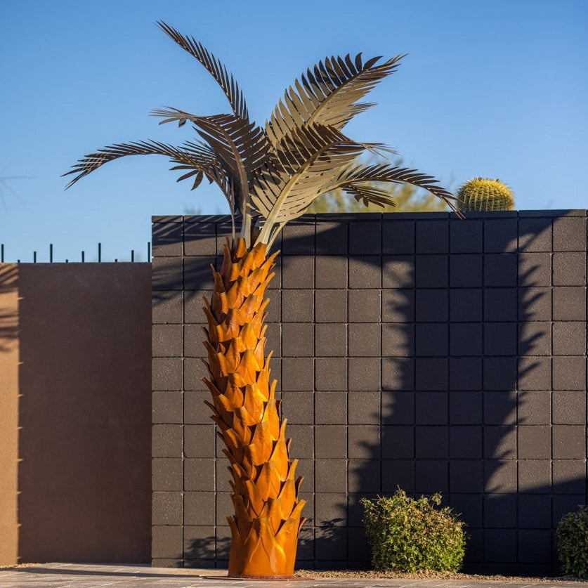 A large metal sunset palm tree, standing 10ft tall, located in a backyard near a pool. The sculpture's intricate design features realistic palm fronds and textured trunk, creating a lifelike appearance.
