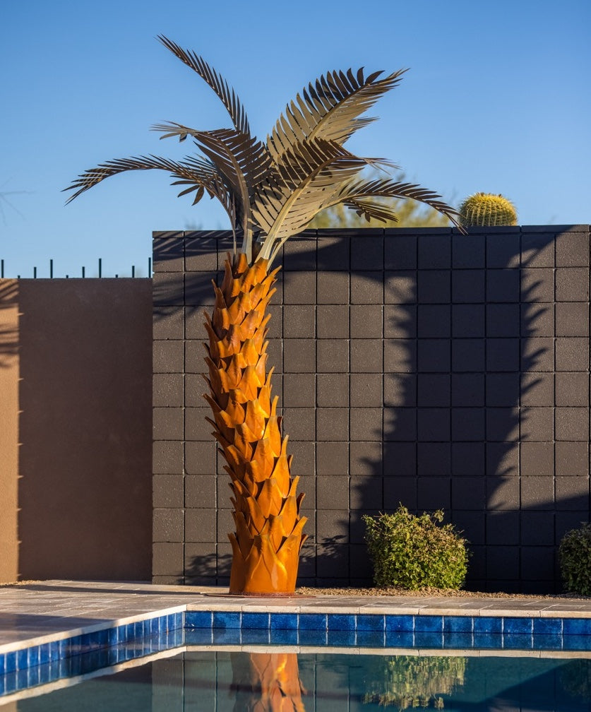 A large metal sunset palm tree, standing 10ft tall, located in a backyard near a pool. The sculpture's intricate design features realistic palm fronds and textured trunk, creating a lifelike appearance.