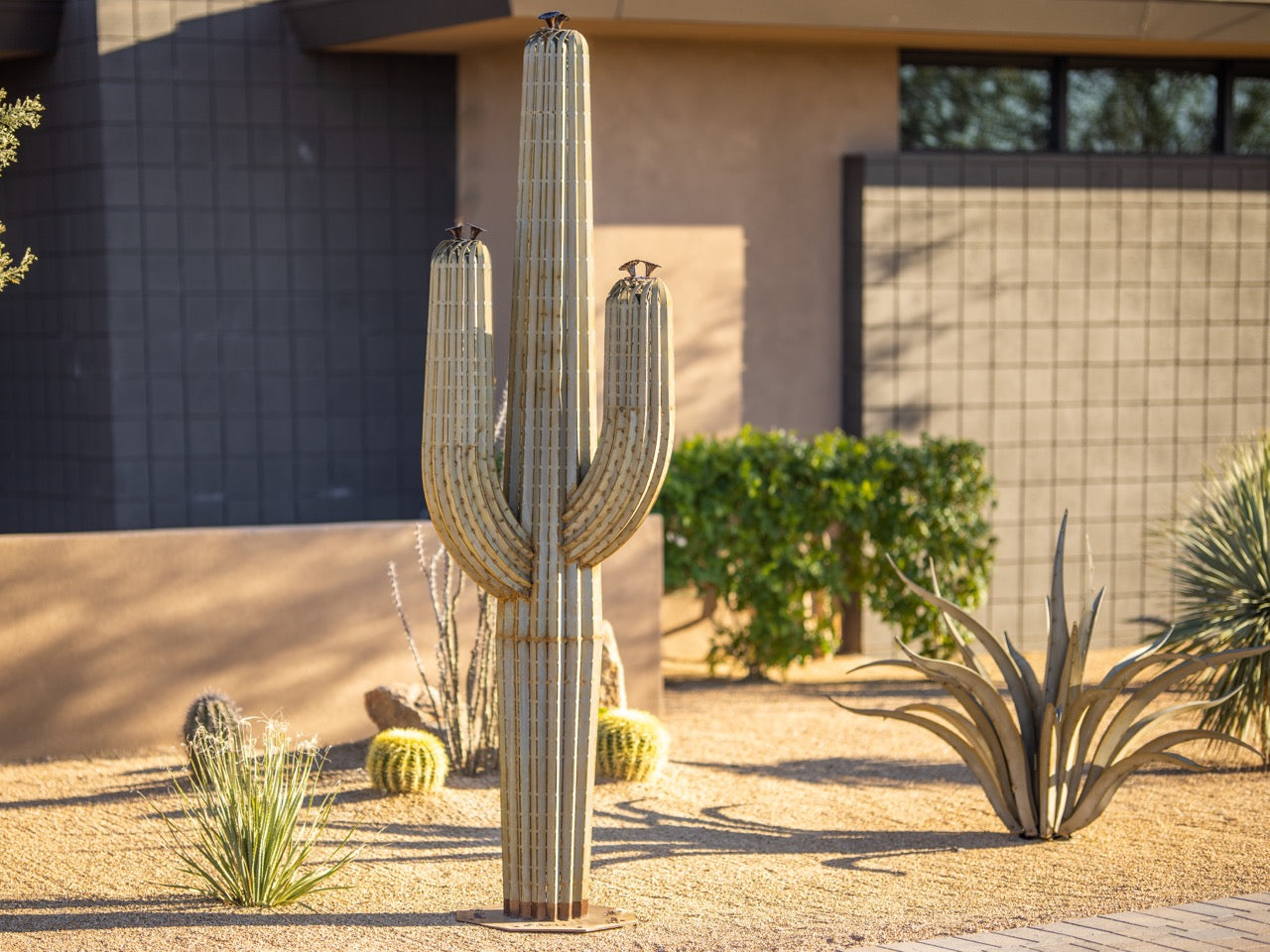 A handcrafted realistic 9ft metal saguaro cactus planted in gravel in the front yard. Surrounded by other plants.