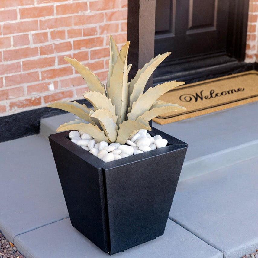Black planter with a plant on a gray mat outside a house entrance.