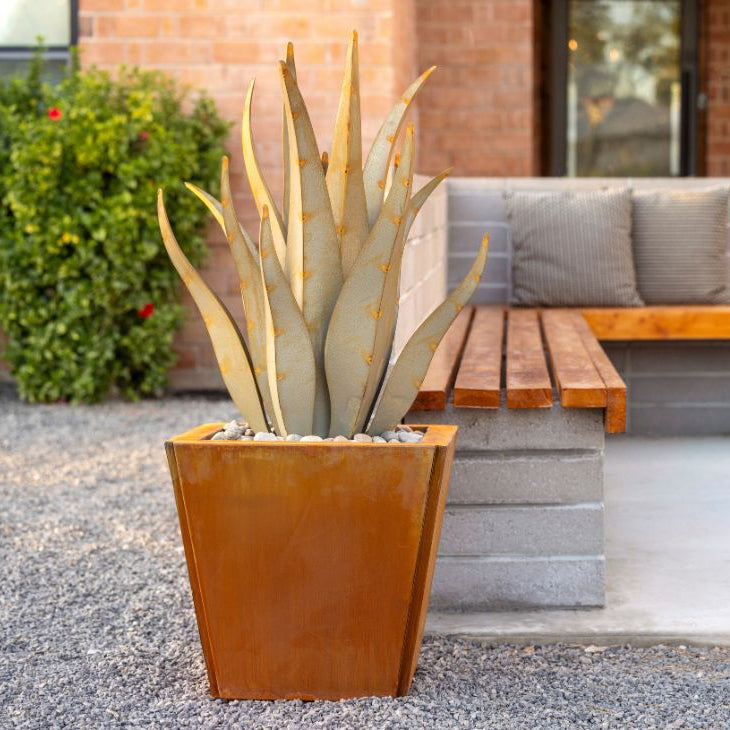 A rectangular brown metal planter with a succulent plant, placed on a gravel surface with a bench and building in the background.