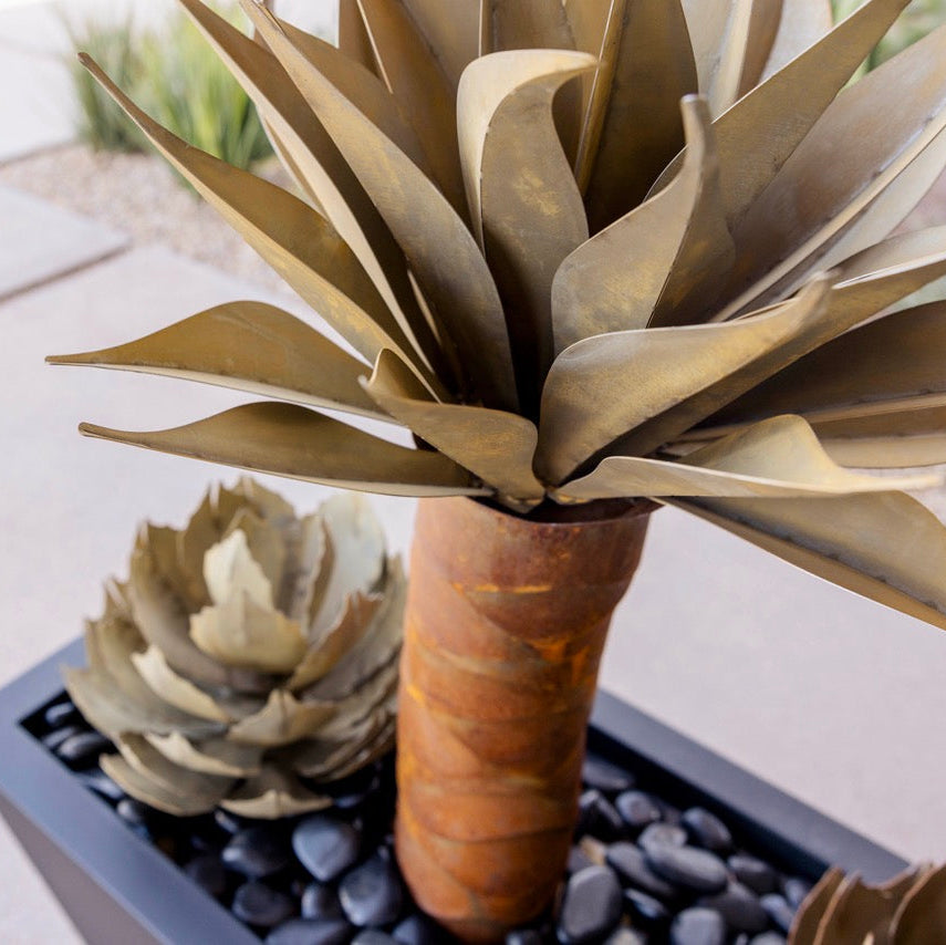 A decorative faux succulent plant with metallic bronze-gray leaves in a tall brown cylindrical pot, shown outdoors in a black rectangular planter filled with dark pebbles.