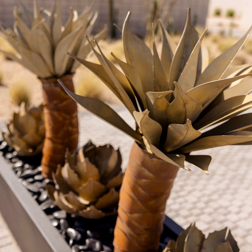 Decorative faux agave plants with metallic gold leaves and brown textured trunks, arranged in a long black rectangular planter filled with black pebbles on a paved outdoor walkway.