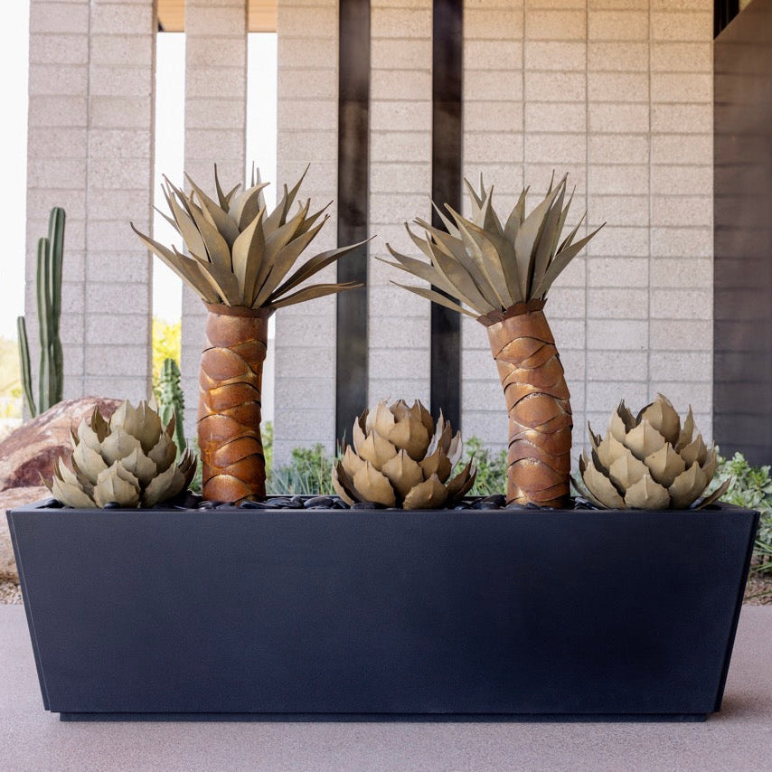 A rectangular black planter box containing four faux agave succulent plants, set outdoors in front of a modern beige stone building.