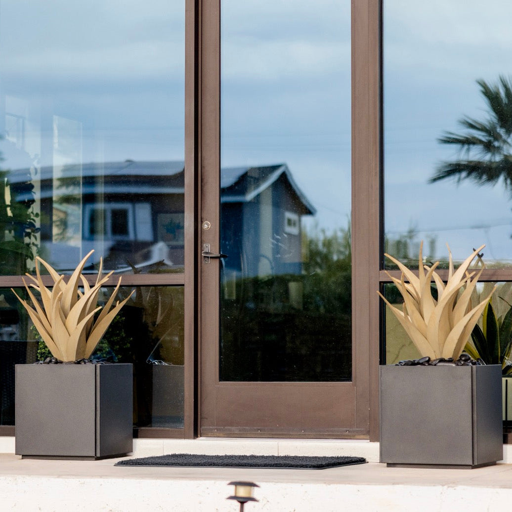 Two steel agaves in planters  in front of a glass door with a view of a house and palm trees.