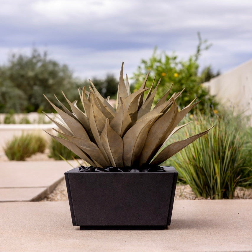A modern black rectangular planter holding a steel agave succulent, set on a concrete patio with a blurred outdoor garden background under a cloudy sky.