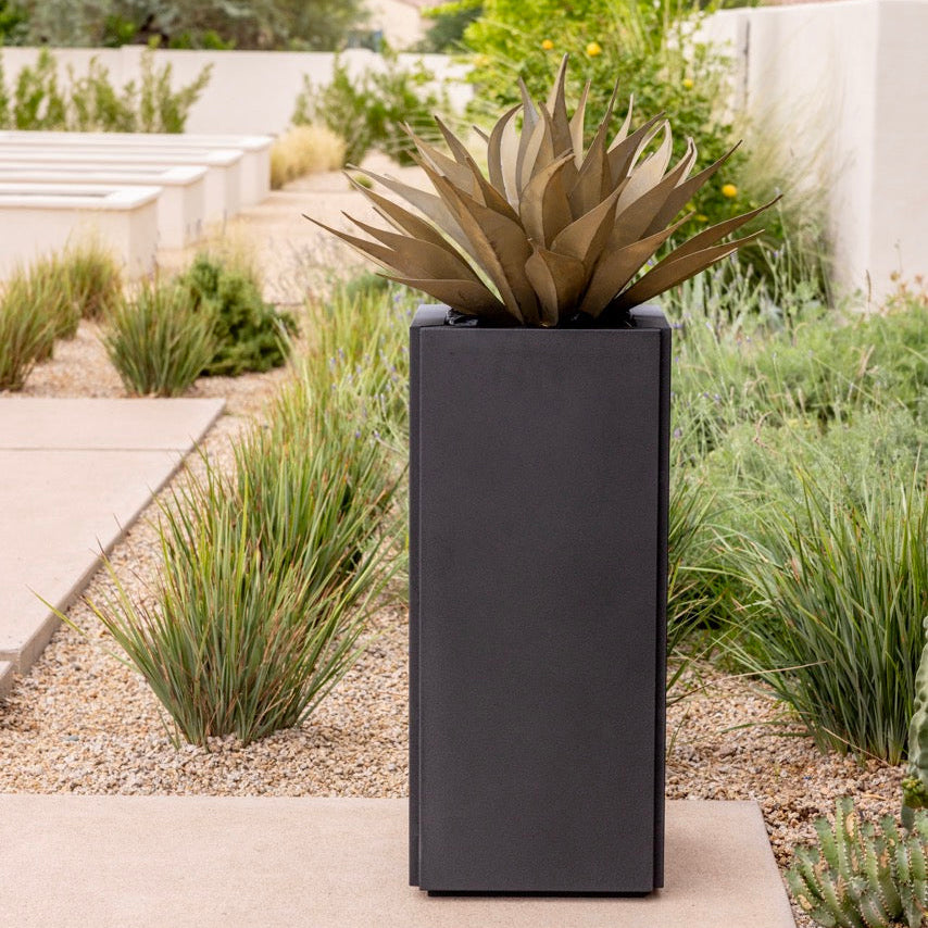 A tall, rectangular matte black planter holding a bronze-toned agave succulent, set on a concrete path in a landscaped garden with desert plants and low walls.