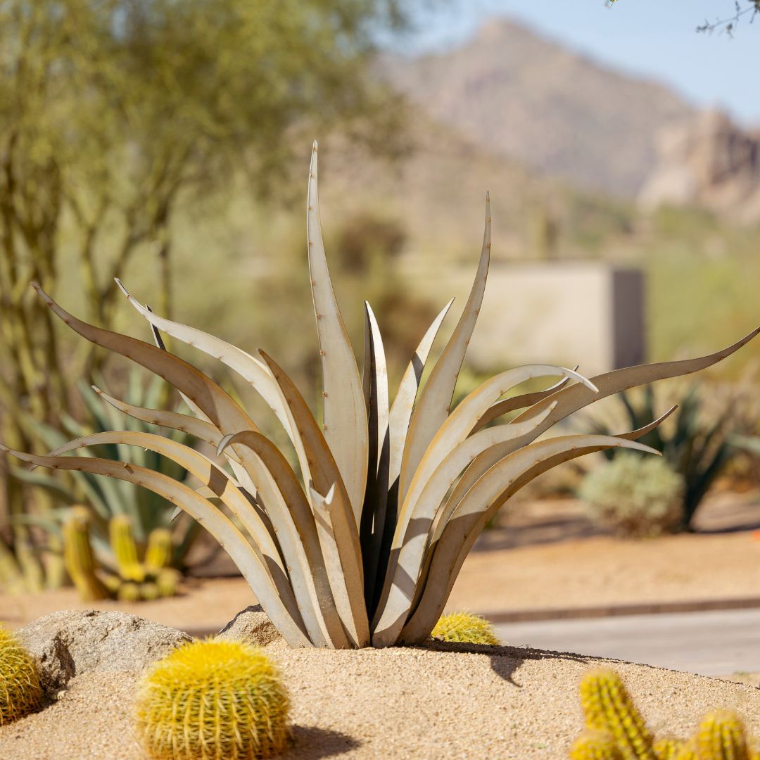 A handcrafted metal sculpture resembling an octopus-shaped agave plant placed in a desert landscape with cacti.