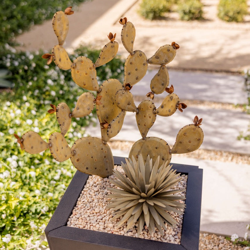 A decorative metal pricked cactus sculpture with multiple flat segments and small spines, displayed in a black square planter alongside a faux agave succulent on white gravel, set outdoors in a garden pathway.