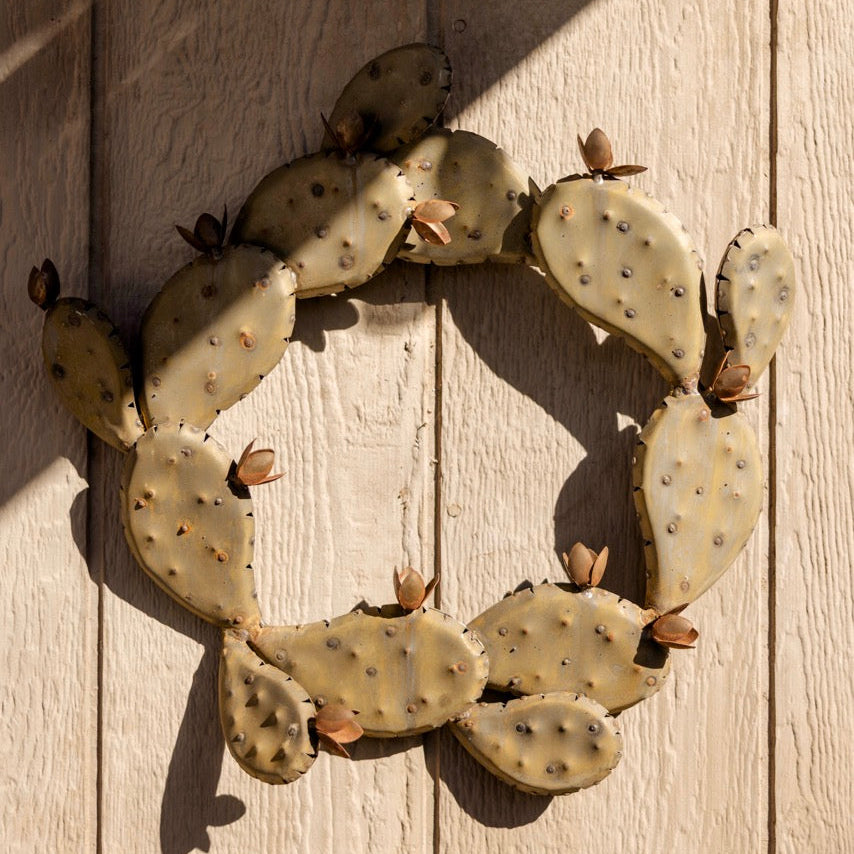 A circular decorative wall piece made of overlapping prickly pear cactus pads with small dried flower buds, mounted on a light wood plank wall under direct sunlight.