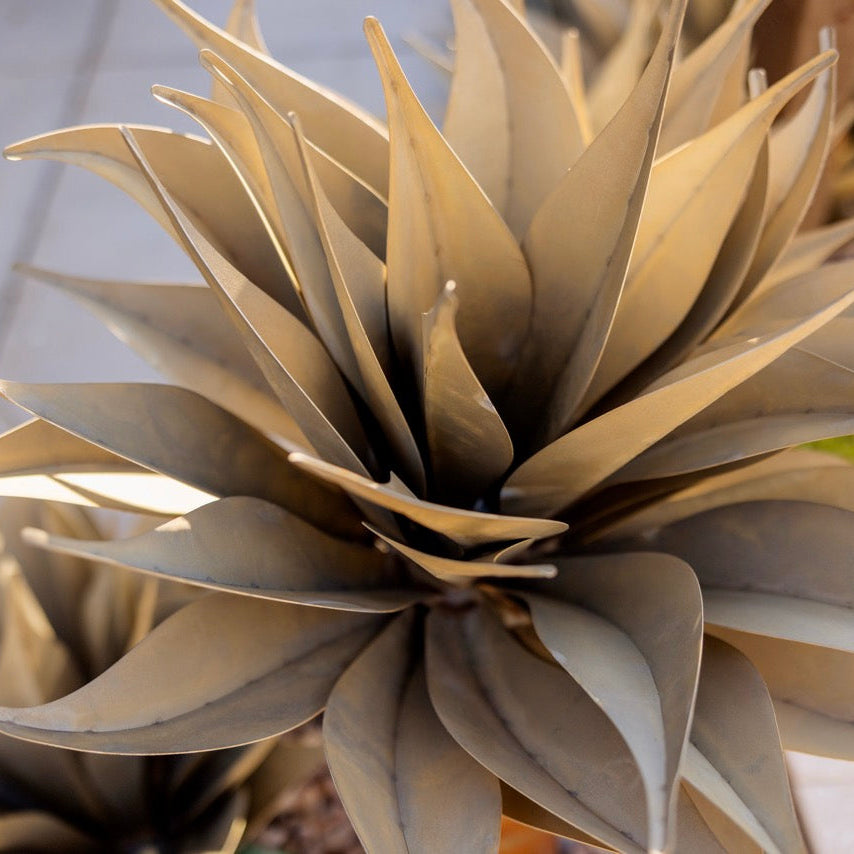 A close-up of a decorative faux agave plant with metallic bronze-gray leaves arranged in a rosette, photographed outdoors on a light stone or concrete surface.