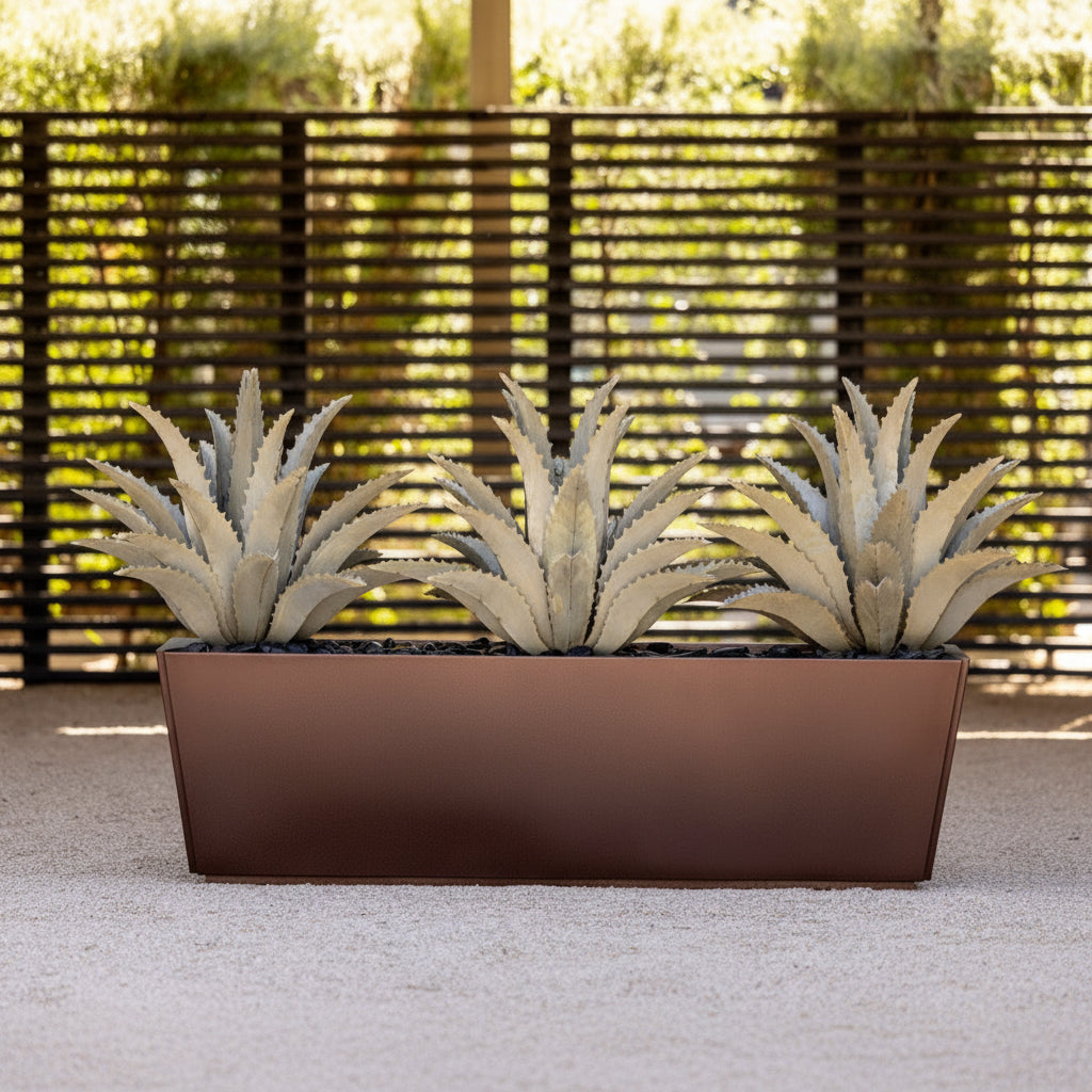 Three steel agave plants in a rectangular bronze planter against a wooden fence.