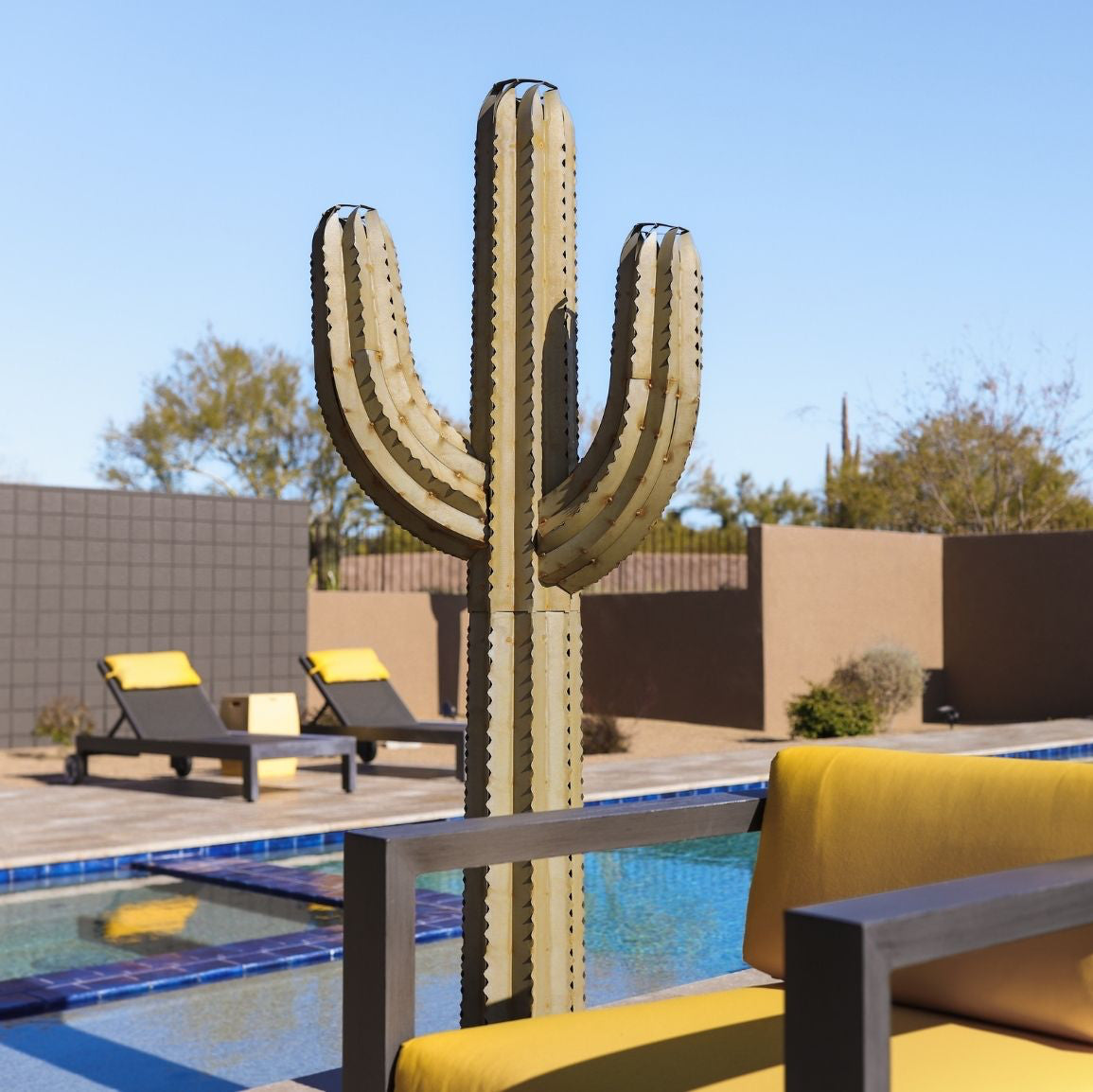 A handcrafted metal torch designed to resemble a Saguaro cactus, with a pool and yellow furniture in the background.