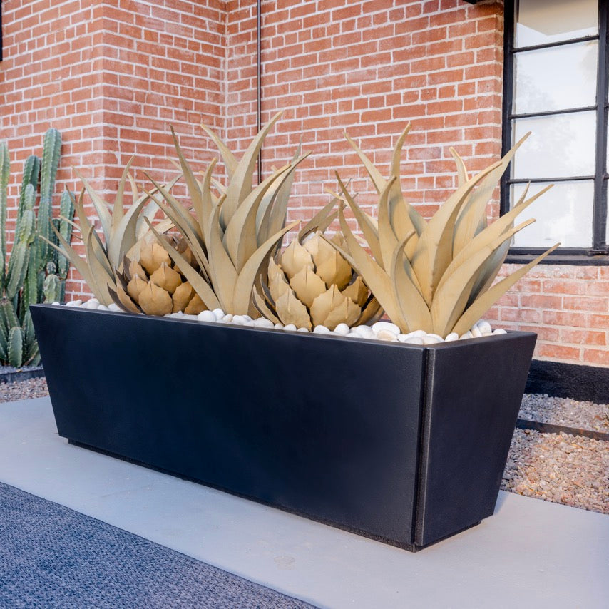 A large rectangular black planter box containing faux agaves, filled with white pebbles, set on a concrete walkway in front of a red brick building.