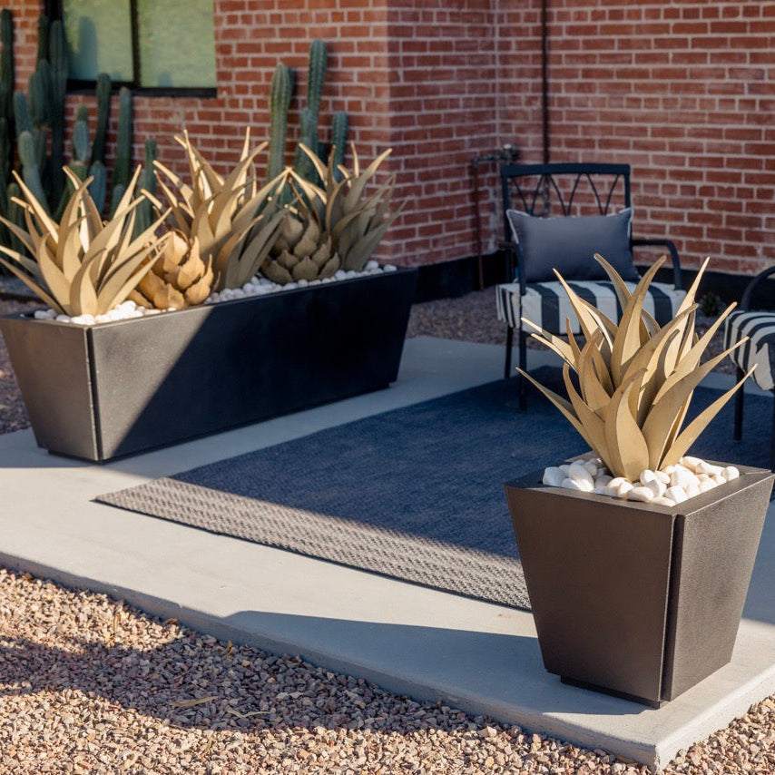 Modern outdoor patio scene with three large dark rectangular planters holding tall spiky faux agave plants in white stones, arranged on a concrete slab beside striped patio chairs and a brick building.