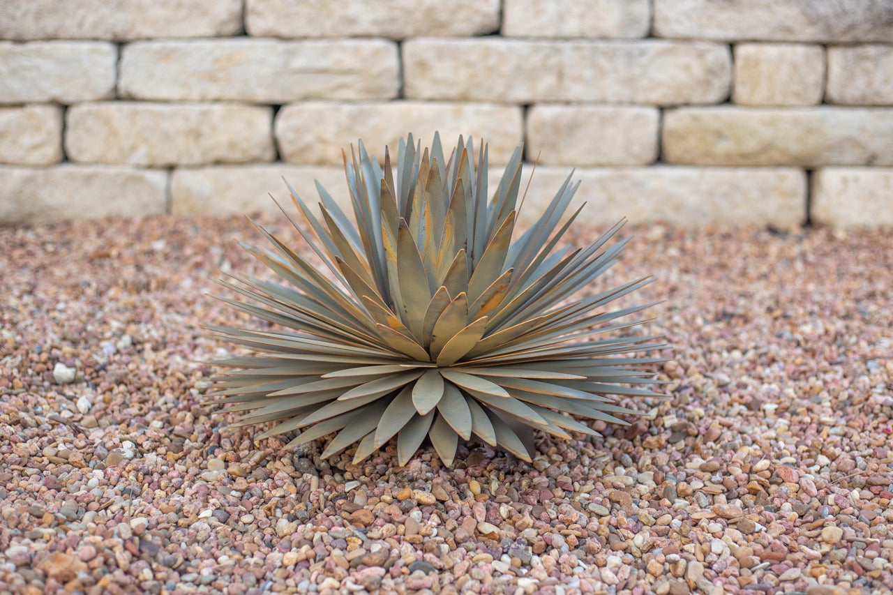 A handcrafted realistic large metal yucca succulent planted in gravel. The succulent is located in front of a white brick wall.