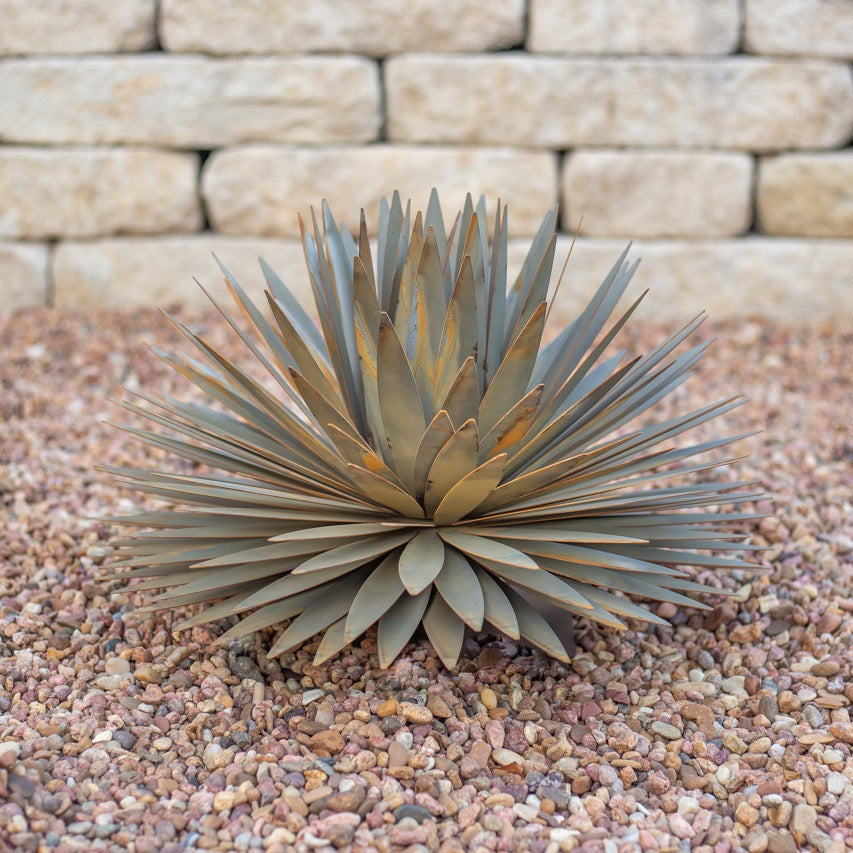 A handcrafted realistic large metal yucca succulent planted in gravel. The succulent is located in front of a white brick wall.