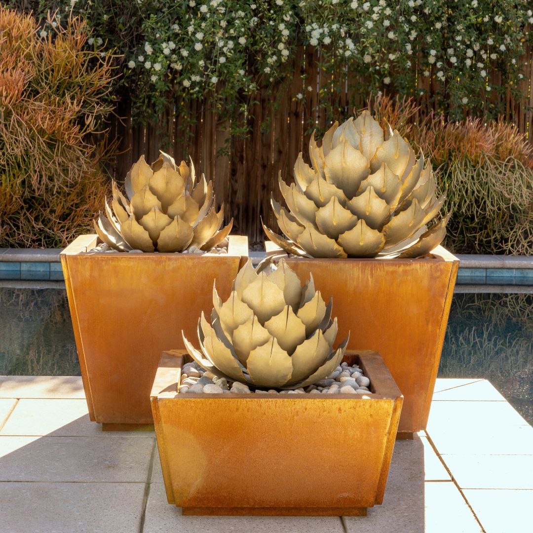 Three metal artichoke agave sculptures inside orange planters, with a garden landscaping in the background. 