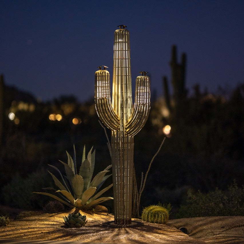 A large metal saguaro cactus illuminated at night in the desert. The sculpture's intricate design features a realistic saguaro body and arms, creating a lifelike appearance.