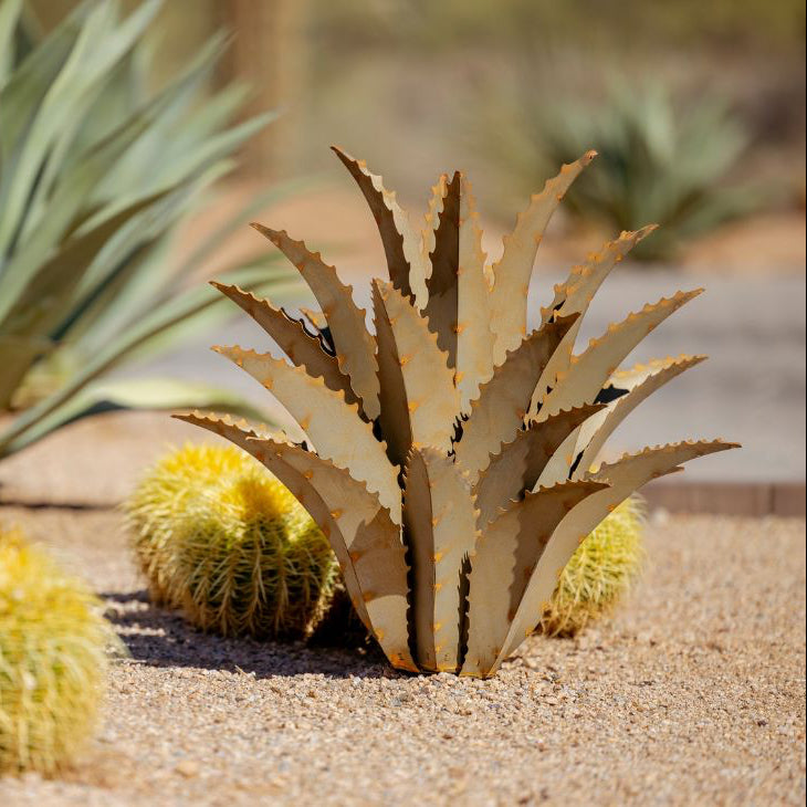A handcrafted metal sculpture resembling a sawtooth agave plant, displayed in a natural setting with other cacti in the background.