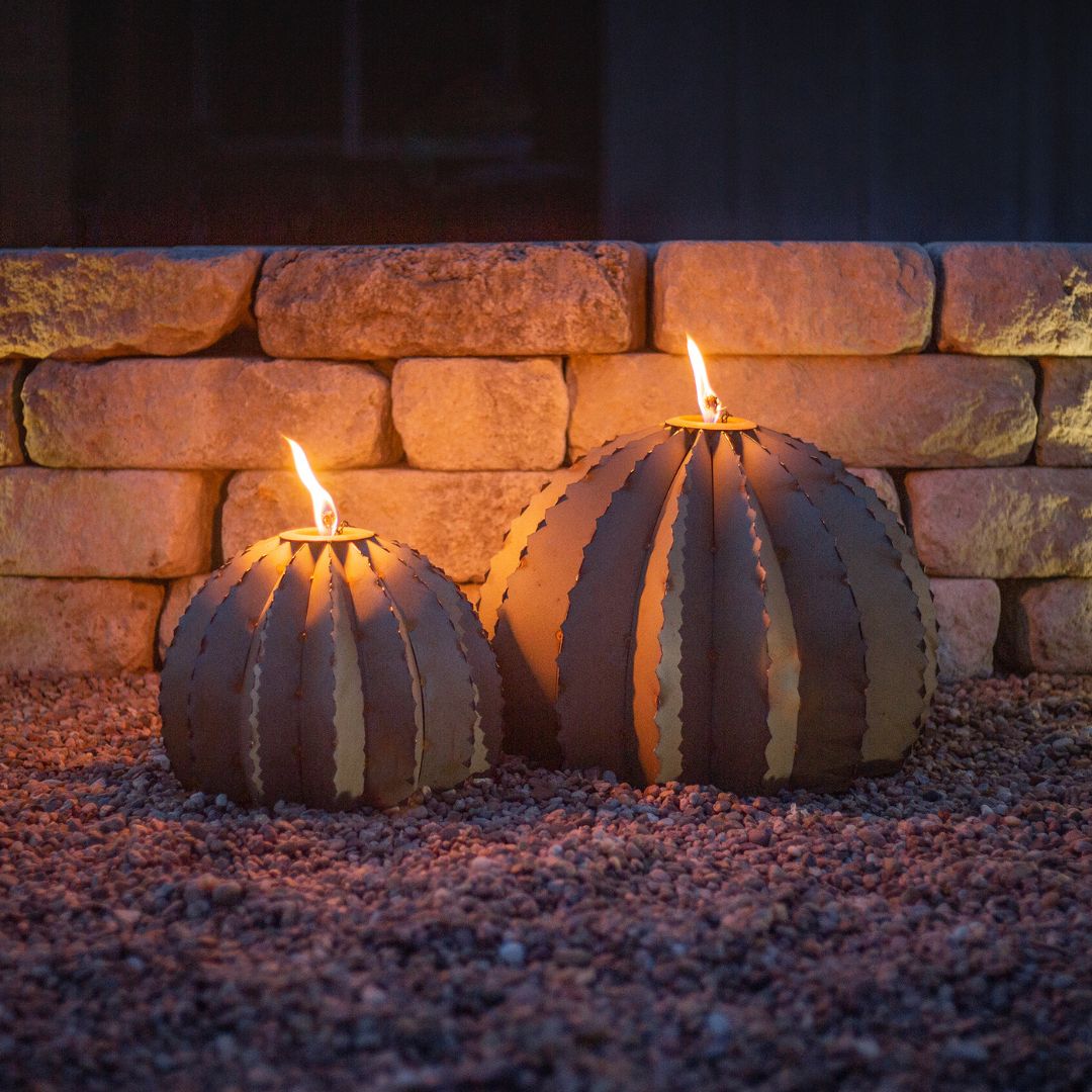 Small and large sized metal barrel cactus torch sculptures lit up, next to eachother on a gravel pavement.