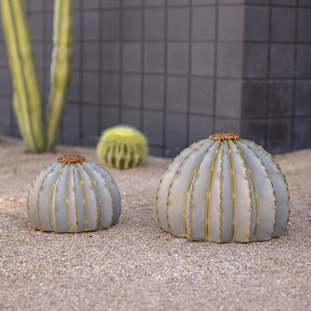 Small and large sized metal barrel cactus torch sculptures on sand next to each other, with a desert landscaping in the background.