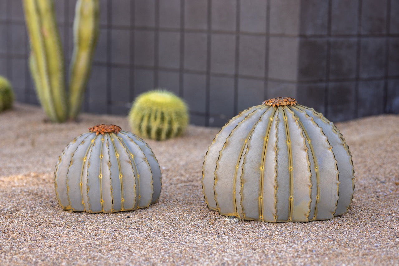 Small and large sized metal barrel cactus torch sculptures on sand next to each other, with a desert landscaping in the background.