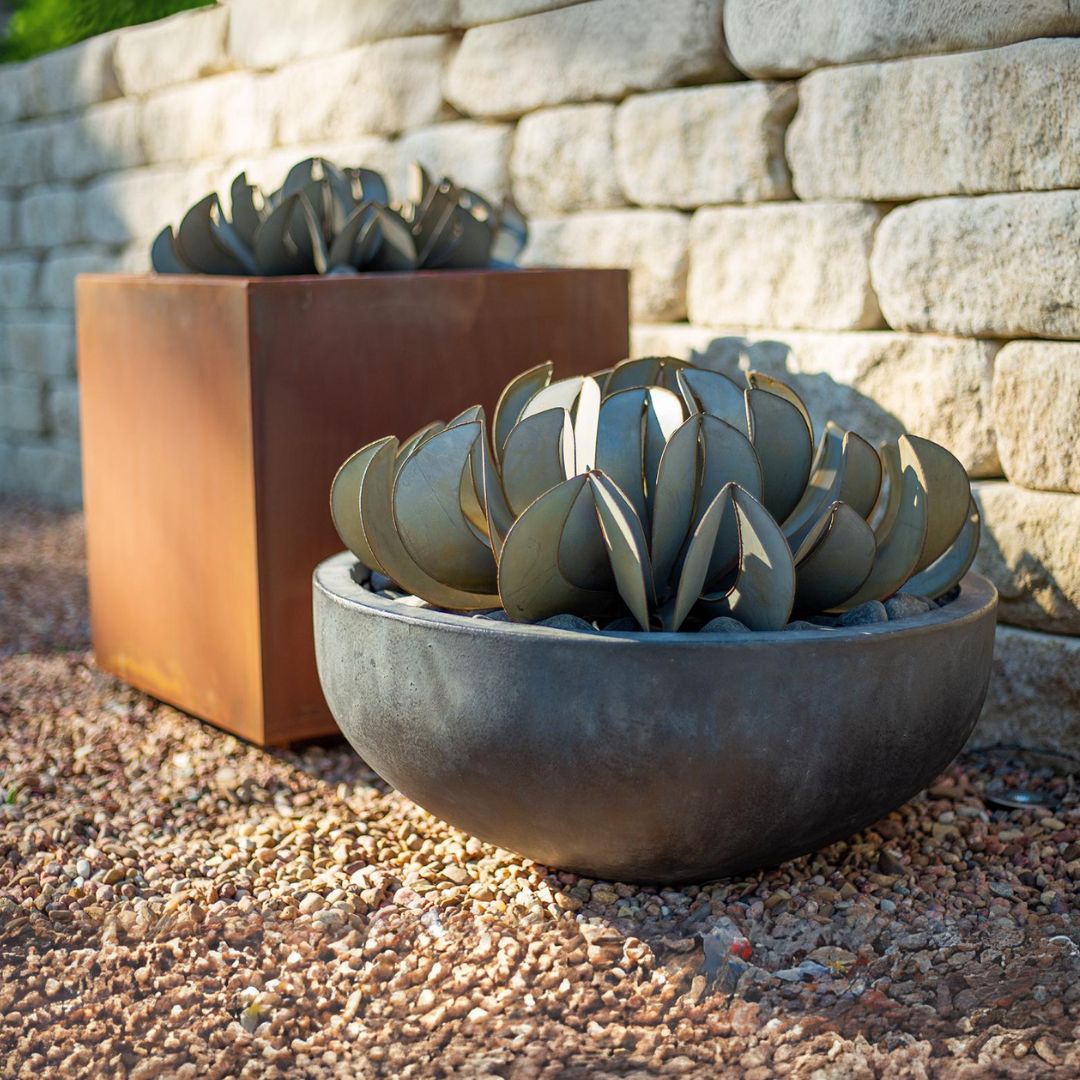 Two metal topsy turvy succulents. One in a cubed orange planter, the other is in a round grey planter. Both located outside next to a brick wall.